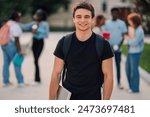 Young adolescent university student boy standing at college campus with laptop under armpit and backpack while smiling at camera. Happy male college student ready for learning posing at campus.