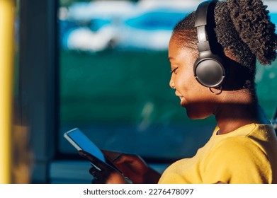 Young adorable african american joyful woman sitting on the bus and using the phone and headphones. Public transportation, technology and people concept. Copy space.