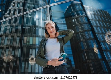 Young adorable blonde swedish woman in headphones I leather jacket and jeans holding phone looking at camera against contemporary building. Student girl in the city, relaxing, listening music.