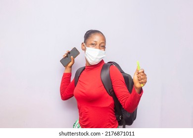 Young Adorable Female Student Smiling In Mask, Holding Mobile Phone And Pen Whilst Carrying Backpack