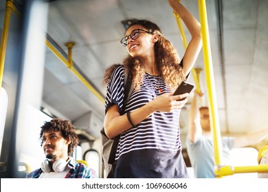 Young adorable joyful woman is standing on the bus using the phone and smiling.