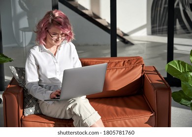 Young Adult 20s Hipster Woman Freelancer Student Distance Worker With Pink Hair Holding Using Laptop Notebook Studying Working From Home Office Online Sitting On Sofa Couch In Living Room Interior.