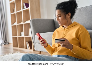 Young adult African American female consumer holding credit card and smartphone sitting on floor at home doing online banking transaction. E commerce virtual shopping, secure mobile banking concept.