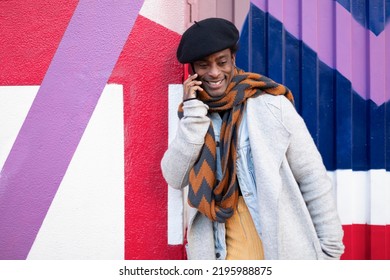 Young Adult African American Man Having A Friendly Phone Conversation. He Is Leaning Against A Colorful Wall.