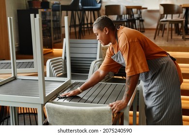 Young Adult African American Woman Starting Workday In Modern Cafe Preparing Tables Turning Them Over And Wiping Dust