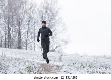 Young, Adult Alone Man In Sport Tracksuit And Hat Running On Park Trail In Winter Day After First Snow. Front View. Empty Place For Motivational, Inspirational Text, Quote, Sayings On Sky Background.