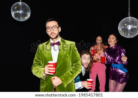young adult arab man standing near funny multiracial friends in colorful clothes at party on black background