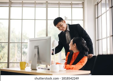 Young Adult Asian Business Man Teaching Work To Woman In A Team With Smile And Empathy. Workplace Table With Computer. Modern Office Background With Window Sunlight.