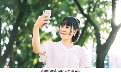 Young Adult Asian Business Single Woman Using Mobile Phone Talking With Friend. Urban Lifestyle Working People. Backgroud With Blur Green Tree.