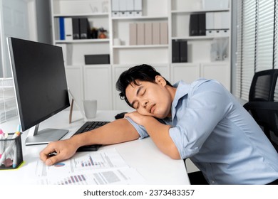 Young adult Asian businessman sleeping on his desk with a computer pc in the office setting background. - Workaholic, boring, exhausted, sleepy business man concept