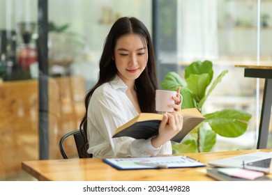 Young Adult Asian Businesswoman Drinking Coffee And Reading Novel For Take A Break From Work, Sitting At Coffee Shop, Relaxing Time