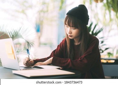 Young Adult Asian College Student Woman Wear Red Sweater And Wool Hat. Using Laptop Study And Work Online At Cafe. University People City Lifestyle At Outside From Home On Winter Day.