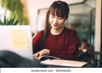 Young Adult Asian College Student Woman Wear Red Sweater. Using Laptop Study And Work Online At Cafe. University People City Lifestyle At Outside From Home On Winter Day.