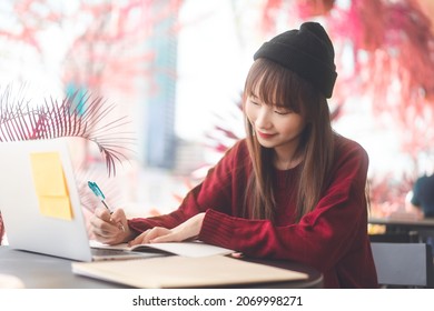 Young Adult Asian College Student Woman Wear Red Sweater. Using Laptop Study And Work Online At Cafe. University People City Lifestyle At Outside From Home On Winter Day.