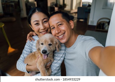 Young Adult Asian Couple Holding A Puppy Taking A Selfie From A Phone With Home Interior In Background. 30s Mature Man And Woman With Dog Pet Taking A Family Photo Shots. - Happy Group Portrait