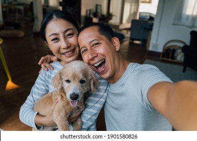 Young Adult Asian Couple Holding A Puppy Taking A Selfie From A Phone With Home Interior In Background. 30s Mature Man And Woman With Dog Pet Taking A Family Photo Shots. - Happy Group Portrait