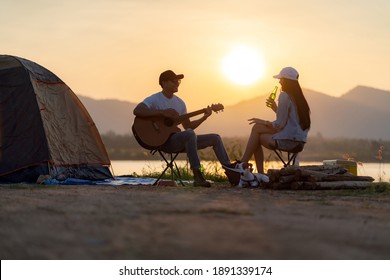 Junge erwachsene asiatische Paare spielen Gitarre und trinken Bier neben ihrem Zeltplatz bei Sonnenuntergang. Campingausflug mit dem Campingplatz-Konzept für Hunde.