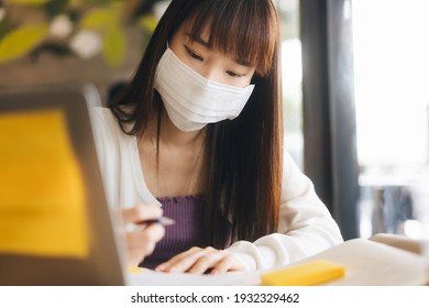 Young Adult Asian Female With Protective Face Mask For Virus Corona Or Covid 19. Work And Study At Cafe New Normal Gig Economy During Pandemic Lifestyle Concept. Indoor On Day.