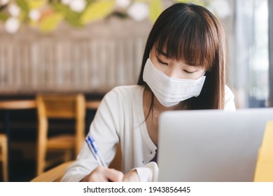 Young Adult Asian Female With Protective Face Mask For Virus Corona Or Covid 19. Work And Study At Cafe New Normal Gig Economy During Pandemic Lifestyle Concept. Indoor On Day.