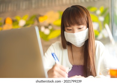 Young Adult Asian Female With Protective Face Mask For Virus Corona Or Covid 19. Work And Study At Cafe New Normal Gig Economy During Pandemic Lifestyle Concept. Indoor On Day.