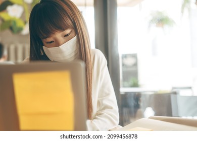 Young Adult Asian Female With Protective Face Mask For Virus Corona Or Covid 19. Work And Study At Cafe New Normal Gig Economy During Pandemic Lifestyle Concept. Indoor On Day.