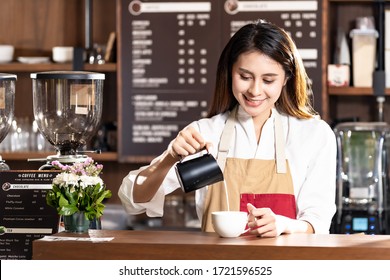 Young adult asian female woman barista pouring fresh milk to prepare latte coffee for customer in cafe bar with her colleague working in background. For small business startup in food industry concept - Powered by Shutterstock