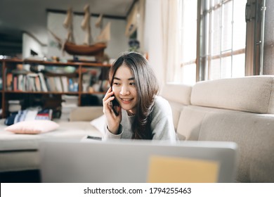 Young Adult Asian Freelancer Business Woman Working Online Via Internet At Home. Using Mobile Phone Call With Customer. Living Room Workspace Background With Window Light.