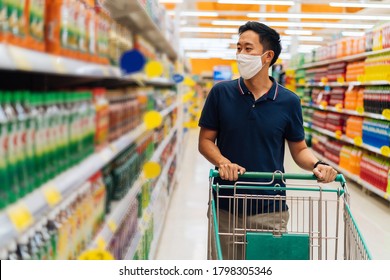 Young Adult Asian Man Wearing A Face Mask While Shopping With Cart Trolley In Grocery Supermarket Store. He's Choosing To Buy Products In The Grocery Store During Covid 19 Crisis In Thailand