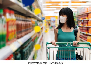 Young Adult Asian Woman Wearing A Face Mask While Shopping With Cart Trolley In Grocery Supermarket Store. She's Choosing To Buy Products In The Grocery Store During Covid 19 Crisis In Thailand