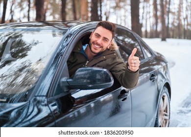 Young adult attractive Caucasian man sits at the wheel of his car sunny winter day shoeing thumbs up gesture. Wintertime road trip. Happy smiling hipster guy sitting in car and looking window. 
