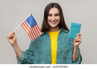 Young adult attractive woman holding united states flag and passport, being happy to move abroad to USA, wearing casual style jacket. Indoor studio shot isolated on gray background. - Shutterstock ID 2127359303