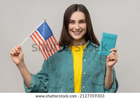 Young adult attractive woman holding united states flag and passport, being happy to move abroad to USA, wearing casual style jacket. Indoor studio shot isolated on gray background.