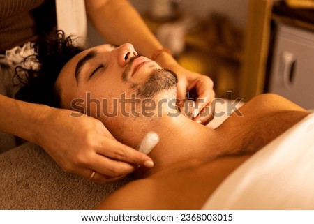 A young adult with a beard is lying on a stretcher in an aesthetic center undergoing a biotherapy session. The beautician places quartz stones on his neck for the elimination of toxins.