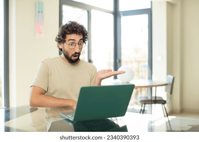 young adult bearded man with a laptop looking surprised and shocked, with jaw dropped holding an object with an open hand on the side