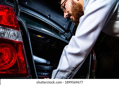 Young Adult, Bearded Man Looking Into A Car Trunk, A Boot At Night.