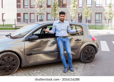 Young adult bearded man with sad expression standing with raised arms and looking at camera, being confused after car accident, broken auto with dents and scratches on side part. Outdoor shot.