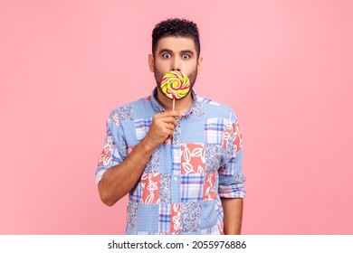 Young adult bearded man wearing blue casual shirt covering his astonished face with round sweet sugary rainbow candy, looking at camera with big eyes. Indoor studio shot isolated on pink background.