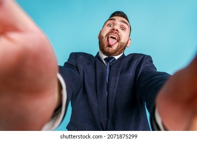 Young adult bearded man wearing dark stylish official style suit, looking at camera with funny expression and showing tongue out POV. Indoor studio shot isolated on blue background.