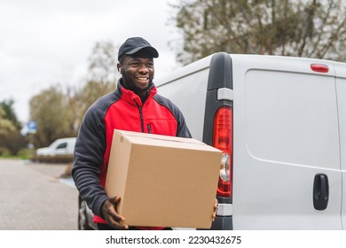 Young adult black delivery guy wearing work uniform smiling carrying cardboard box package unloaded from white van. Horizontal outdoor shot. High quality photo