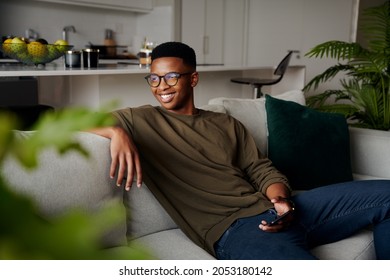 Young adult black male relaxing on sofa holding cellphone in modern apartment
