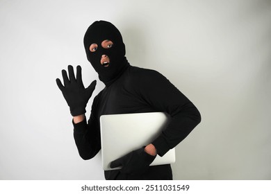 Young Adult black masked robber is holding silver colored laptop. Running, fleeing and escaping pose. Concept for hacking, scamming, phishing. - Powered by Shutterstock