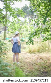 Young Adult Blonde Curvy Woman Stands In The Middle Of The Forest On A Sunny Day Of Summer. She Is Wearing A Jeans Dress, White Shirt And A Hat.