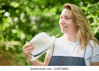 Young Adult Blonde Curvy Woman Stands In The Middle Of The Forest On A Sunny Day Of Summer. She Is Wearing A Jeans Dress, White Shirt And A Panama Hat.