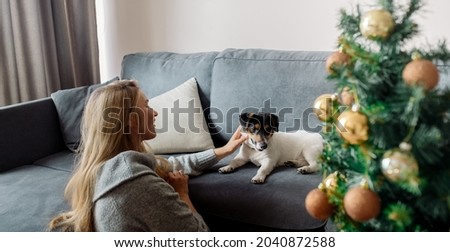 Young adult blonde woman sitting at home on the sofa next to christmas tree and playing with dog 