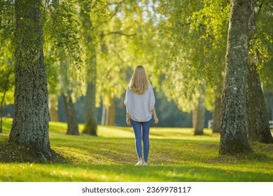 Young adult blonde woman slowly walking on green grass at city park in beautiful warm sunny autumn day. Spending time alone and enjoying freedom at nature. Peaceful atmosphere. Back view. - Powered by Shutterstock