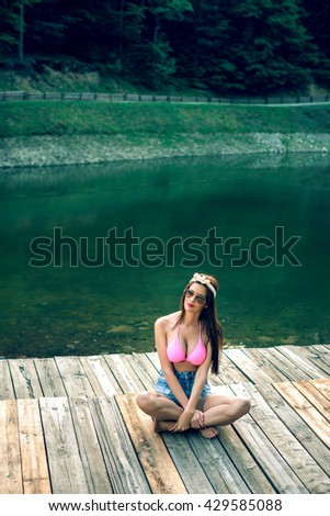 Young adult brunette female posing on wooden pier of small mountain lake wearing retro styled clothes.