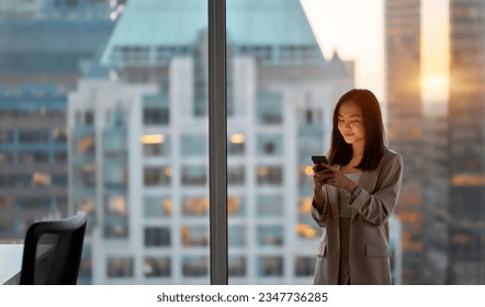 Young adult busy Asian business woman executive using mobile cell phone technology in office. Professional lady businesswoman working on digital smartphone device with city view from window. - Powered by Shutterstock