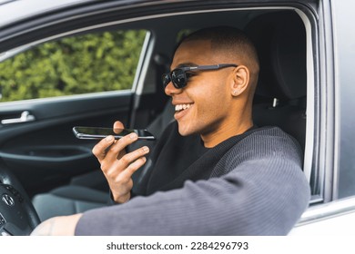 Young adult casually dressed Black man in sunglasses sitting behind a wheel and talking via loudspeaker on his phone. Portrait shot through driver's window. High quality photo - Powered by Shutterstock