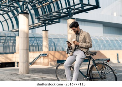 A young adult Caucasian businessman in smart casual attire leans on his bicycle, engaged with his smartphone, enjoying a takeaway coffee outside a modern urban structure. - Powered by Shutterstock