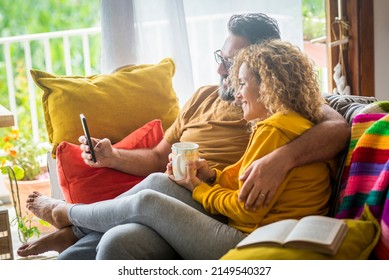 Young Adult Caucasian Couple Enjoy Life Together At Home Doing Video Call At Friends Sitting On The Sofa And Enjoying Wireless Connection. Modern People Use Mobile Cellular On The Couch